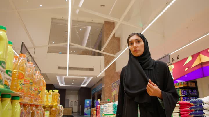 The concept of smart shopping, an Arab Gulf Emirati woman wearing a hijab and an abaya stands inside the supermarket, thinking and carefully selecting food products from the shelves, buying daily necessities, making purchasing decisions, shopping in modern stores.