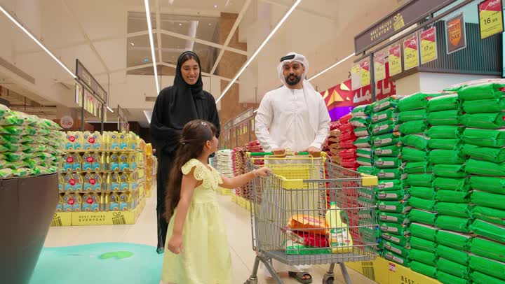 عائلة عربية خليجية إماراتية ترتدي الزي التقليدي داخل متجر مواد غذائية، اختيار وشراء الاحتياجات اليومية، التسوق العائلي في السوبر ماركت ، بنتتشير بيدها إلى المنتجات على الرفوف أثناء التسوق برفقة والديها، مفهوم التسوق الأسري