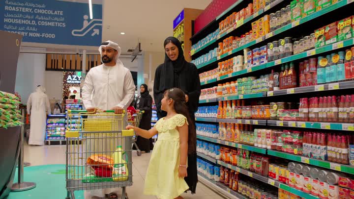 اختيار وشراء الاحتياجات اليومية، التسوق العائلي في السوبر ماركت ، بنت تشير بيدها إلى المنتجات على الرفوف أثناء التسوق برفقة والديها، عائلة عربية خليجية إماراتية ترتدي الزي التقليدي داخل متجر مواد غذائية، مفهوم التسوق الأسري