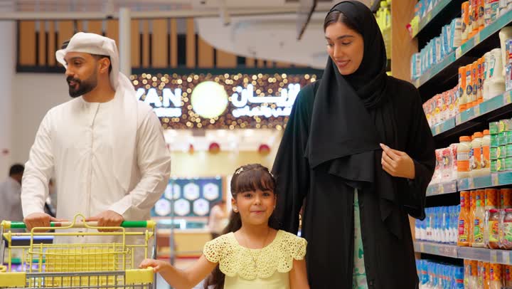 التسوق العائلي في السوبر ماركت ، بنتتشير بيدها إلى المنتجات على الرفوف أثناء التسوق برفقة والديها، عائلة عربية خليجية إماراتية ترتدي الزي التقليدي داخل متجر مواد غذائية، اختيار وشراء الاحتياجات اليومية، مفهوم التسوق الأسري