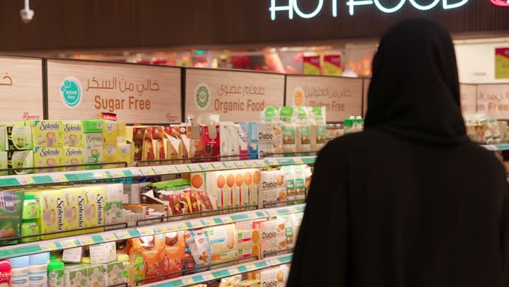 مقارنة المنتجات وقراءة التفاصيل قبل الشراء، تصوير من الخلف لامرأة عربية خليجية اماراتية محجبة تسير داخل السوبر ماركت برفقة زوجها و ابنتها، تفقد المنتجات الغذائية و مفهوم التسوق عائلي، اختيار وشراء المواد الغذائية