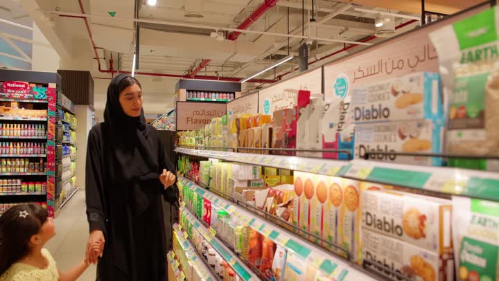 مقارنة المنتجات والتسوق العائلي، عائلة عربية خليجية اماراتية ترتدي الزي التقليدي تتسوق داخل سوبر ماركت، امراه اماراتية تقوم بقراءة تفاصيل العبوة قبل الشراء، اختيار وشراء المواد الغذائية