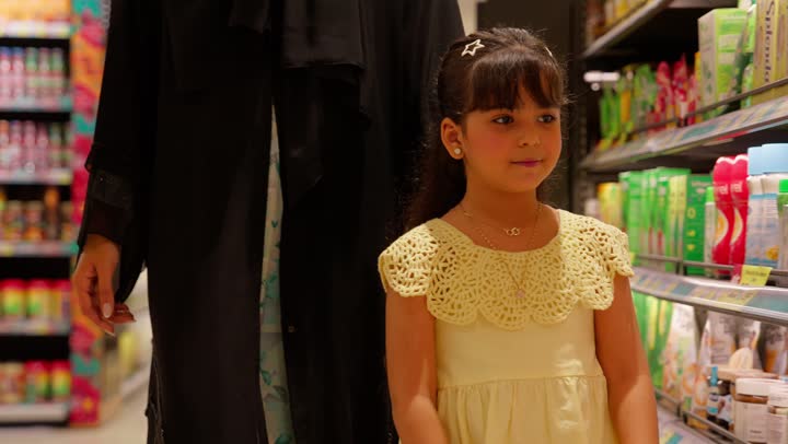 مفهوم التسوق العائلي وشراء الاحتياجات المنزلي، بنت عربية خليجية اماراتية ترتدي الفستان تتسوق داخل سوبرماركت برفقة والديها ، اختيار المنتجات الغذائية من رفوف المتجر، ايماءات السعادة و السرور