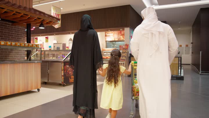 التعاون في شراء السلع والمواد الغذائيه المختلفة في المتجر، مساعدة وتعليم الأبناء الشراء من الماركت، تصوير من الخلف لزوجان عربيان خليجيان اماراتيان يرتديان الزي التقليدي يقومان بقضاء أوقات ممتعة بالتسوق واختيار اللوازم المنزليه برفقه ابنتهم، التمتع بالتخفيضات والعروض والخصومات على المنتجات