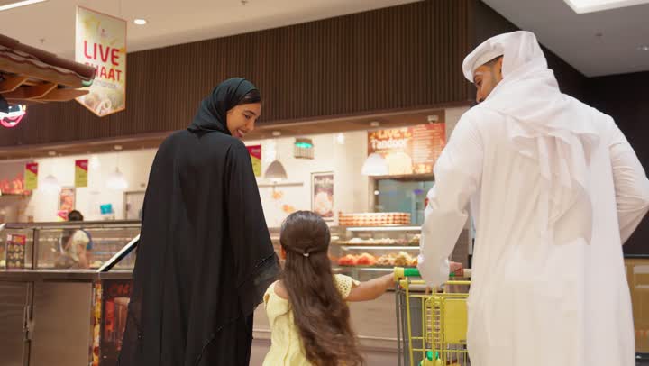 مساعدة وتعليم الأبناء الشراء من الماركت، التعاون في شراء السلع والمواد الغذائيه المختلفة في المتجر، تصوير من الخلف لزوجان عربيان خليجيان اماراتيان يرتديان الزي التقليدي يقومان بقضاء أوقات ممتعة بالتسوق واختيار اللوازم المنزليه برفقه ابنتهم، التمتع بالتخفيضات والعروض والخصومات على المنتجات