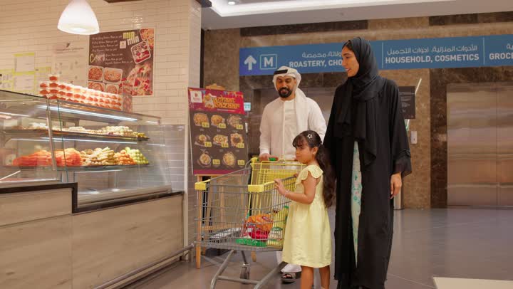 عائلة عربية خليجية اماراتية ترتدي الزي التقليدي تقوم بشراء المواد الغذائية اللازمة داخل السوبرماركت ، شراء الاحتياجات المنزلية ، سلة التسوق الشرائية، ايماءات السعادة و السرور