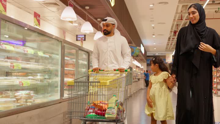 تعليم البنات مفهوم التسوق وحسن الإختيار، شراء الحلويات والكيك، الاشارة الى شيء ما، عائلة عربية خليجيه اماراتية تتسوق داخل قسم المخبوزات في السوبر ماركت ، بنت ترتدي الفستان تشير إلى الحلويات داخل ثلاجة العرض الزجاجية برفقة والديها
