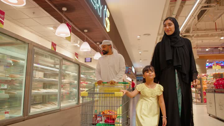 قضاء وقت ممتع مع الابناء، شراء الحلويات والكيك، الاشارة الى شيء ما، مفهوم التسوق العائلي، عائلة عربية خليجيه اماراتية تتسوق داخل قسم المخبوزات في السوبر ماركت ، بنت ترتدي الفستان تشير إلى الحلويات داخل ثلاجة العرض الزجاجية برفقة والديها