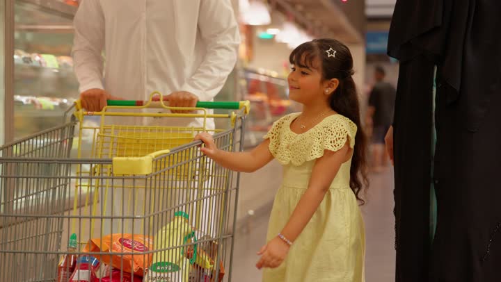 Buying sweets and cakes, pointing to something, the concept of family shopping, an Emirati Gulf Arab family shopping in the bakery section of the supermarket, a girl wearing a dress pointing to the sweets inside the glass display fridge with her parents, gestures of happiness and joy.