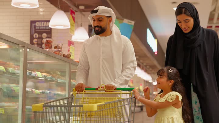 الاشارة الى شيء ما، مفهوم التسوق العائلي، عائلة عربية خليجيه اماراتية تتسوق داخل قسم المخبوزات في السوبر ماركت ، بنت ترتدي الفستان تشير إلى الحلويات داخل ثلاجة العرض الزجاجية برفقة والديها، شراء الحلويات والكيك، ايماءات السعادة و السرور