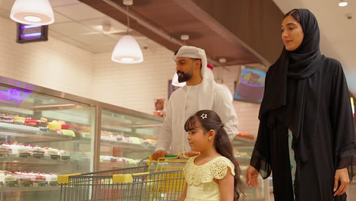 مفهوم التسوق العائلي، عائلة عربية خليجيه اماراتية تتسوق داخل قسم المخبوزات في السوبر ماركت ، بنت ترتدي الفستان تشير إلى الحلويات داخل ثلاجة العرض الزجاجية برفقة والديها، شراء الحلويات والكيك
