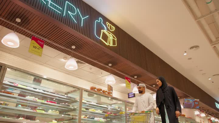 ايماءات الفرح والسعادة بشراء المستلزمات، رجل عربي خليجي اماراتي يرتدي الكندورة و الغترة يمسك بيده سلة التسوق ويقوم بالتجول برفقة زوجته في المتجر، إمراة اماراتية مبتسمة ترتدي العباءة السوداء تشارك زوجها في شراء الحاجيات، قضاء وقت ممتع في السوبرماركت