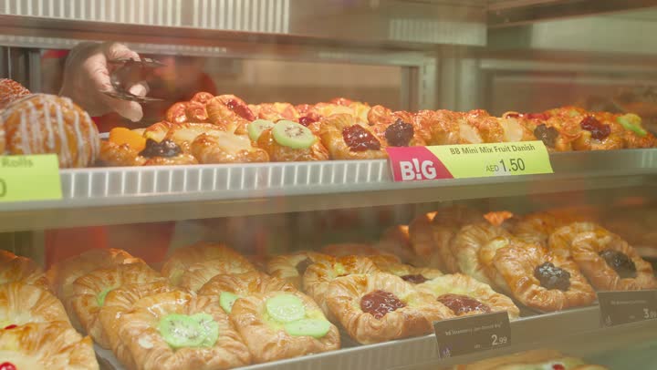 كرواسون ومعجنات متنوعة داخل ثلاجة زجاجية، مفهوم شراء المعجنات والوجبات الخفيفة داخل السوبرماركت، تصوير مقرب ليد عامل يقوم باختيارالمخبوزات باستخدام ملقط