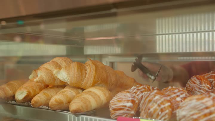 The concept of buying pastries and snacks inside the supermarket, croissants and various pastries inside a glass refrigerator, a close-up shot of a worker's hand selecting baked goods using tongs.