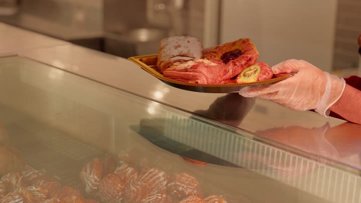 شراء الحلويات والوجبات الخفيفة، مفهوم خدمة العملاء داخل المتجر، تصوير مقرب ليد عامل عربي خليجي اماراتي داخل قسم المخبوزات في السوبر ماركت يقوم بتقديم طبق حلويات طازجة لأحد الزبائن، عرض المنتجات داخل ثلاجة زجاجية