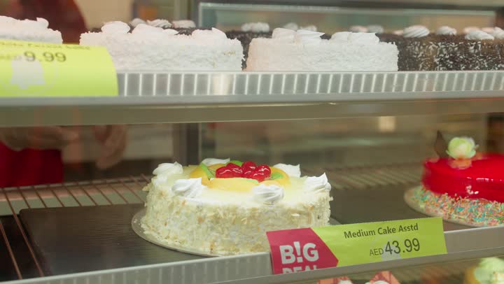 تصوير مقرب ليد عامل عربي خليجي اماراتي داخل قسم الحلويات في سوبر ماركت حديث تقوم بتسليم كعكة مزينة بالكريمة إلى الزبون، خدمة العملاء داخل قسم الديلي، شراء الحلويات والكيك، منتجات مخبوزة طازجة، مفهوم التسوق داخل المتجر