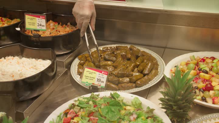 عرض مأكولات عربية شعبية داخل ثلاجة العرض، تصوير مقرب ليد عامل عربي خليجي اماراتي داخل قسم الأطعمة الجاهزة في سوبر ماركت يقوم بتقديم ورق العنب (الدولمة) باستخدام ملقط معدني، وجبات جاهزة ومأكولات مطهية، مفهوم التسوق وشراء الطعام