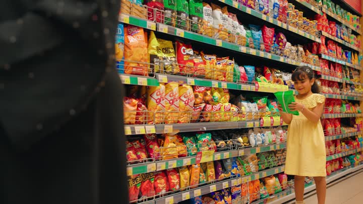 اختيار المنتجات والوجبات الخفيفة، طفلة عربية خليجيه اماراتية ترتدي فستان أصفر تمسك كيس تسالي أثناء التسوق داخل السوبر ماركت برفقة والدتها، مفهوم التسوق العائلي، ايماءات السعادة و السرور