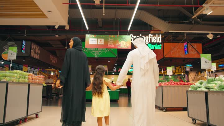 الاستفاده من التخفيضات والعروض على المنتجات، تصوير من الخلف لعائلة عربية خليجيه اماراتية ترتدي الزي التقليدي تتسوق داخل قسم الخضروات والفواكه في السوبر ماركت، أجواء عائلية سعيدة، مفهوم التسوق اليومي، شراء المواد الغذائية، أسلوب حياة صحي، الاهتمام بالغذاء الطازج