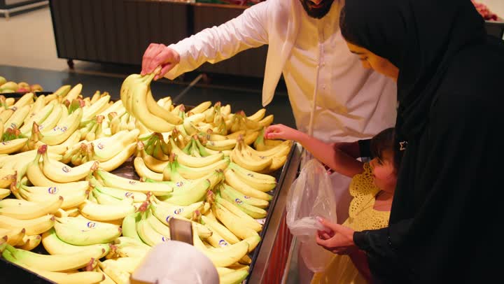 عائلة عربية خليجيه اماراتية ترتدي الزي التقليدي تقف داخل قسم الفواكه في السوبرماركت ، اختيار ثمار الموز الطازجة، التعاون الأسري أثناء التسوق، أجواء عائلية دافئة، اختيار الفواكه الطازجة، الاهتمام بالتغذية الصحية، مفهوم الأسرة السعيدة