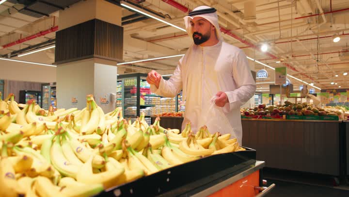 The concept of daily shopping, high-quality Emirati agricultural crops, providing essential nutrients for body health, an Arab Gulf Emirati man wearing a kandura and ghutrah is selecting fresh bananas and putting them in a plastic bag while shopping.