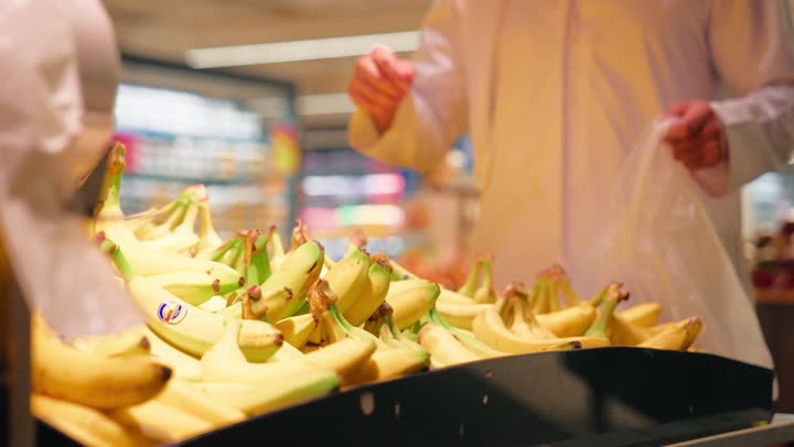 المحاصيل الزراعية الاماراتية ذات الجودة العالية، توفير العناصر الغذائية الضرورية لصحة الجسم، تصوير مقرب لرجل عربي خليجي اماراتي يرتدي الكندورة يقوم باختيار الموز الطازج ووضعه داخل كيس بلاستيكي أثناء التسوق، مفهوم التسوق اليومي