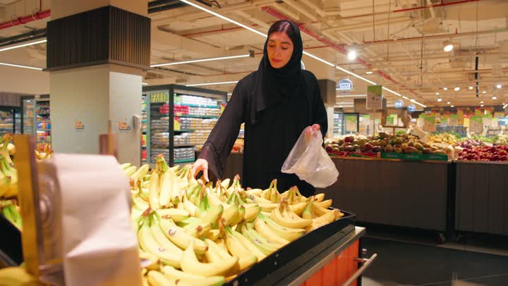 قسم الفواكه داخل السوبر ماركت، شراء المنتجات الغذائية الطازجة، امرأة عربية خليجية اماراتية ترتدي العباءة والحجاب تقوم باختيار الموز الطازج ووضعه داخل كيس بلاستيكي أثناء التسوق، مفهوم التسوق اليومي، أسلوب حياة صحي، الاهتمام بالتغذية الصحية