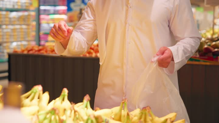 Providing essential nutrients for the body's health, a close-up shot of an Emirati Gulf Arab man wearing a kandura selecting fresh bananas and placing them in a plastic bag while shopping, buying groceries and fresh products from the supermarket, the concept of daily shopping, a healthy lifestyle.