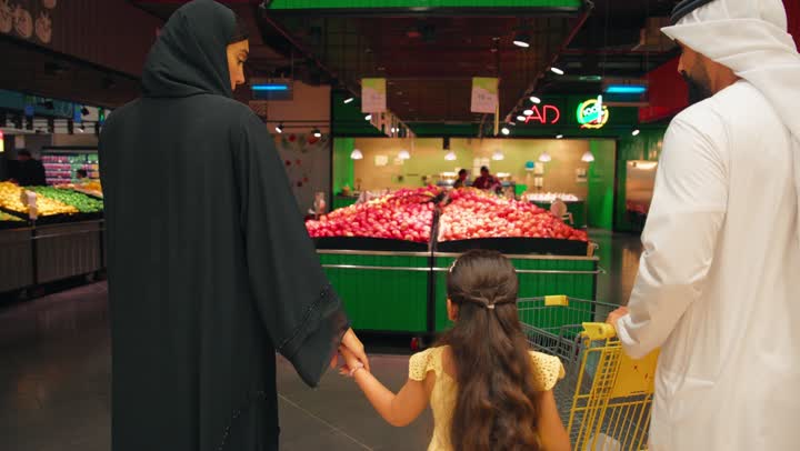 An Emirati Gulf Arab family dressed in traditional attire is shopping inside a supermarket, selecting fresh red apples from the fruits and vegetables section. There is a happy family atmosphere, shopping and buying groceries, a healthy lifestyle, and the concept of family and family bonding.