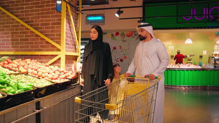 مفهوم التسوق العائلي، عائلة عربية خليجية اماراتية داخل سوبرماركت في قسم الخضروات،بنت تقوم  باختيار ثمرة ملفوف (كرنب) من الخضار الطازج، شراء المنتجات الغذائية الطازجة والاهتمام بالتغذية الصحية