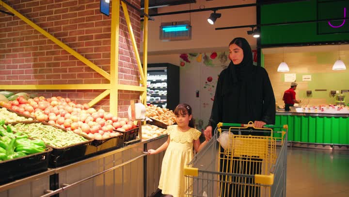 مفهوم الاهتمام بالغذاء الصحي وقضاء وقت ممتع مع الابناء داخل السوبر ماركت، امرأة عربية خليجية اماراتية ترتدي العباءة والحجاب تمسك بيد ابنتها أثناء التسوق في قسم الخضار والفواكه الطازجة، الاشارة إلى المنتجات المعروضة، التسوق العائلي وشراء الاحتياجات اليومية