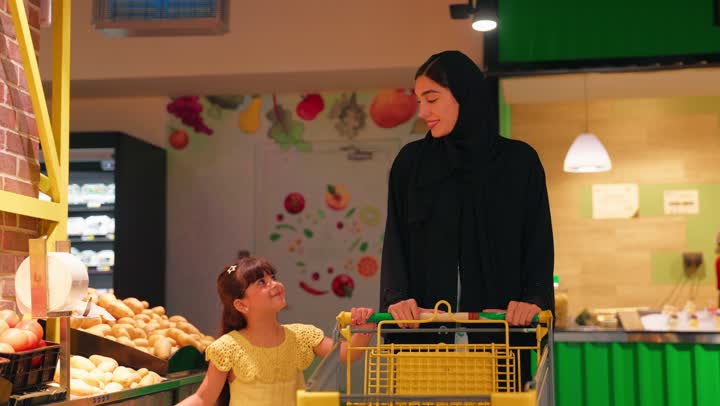Family shopping and attention to healthy nutrition and product quality, an Emirati Gulf Arab woman wearing an abaya and hijab is accompanied by her daughter as they push a shopping cart in the fruits and vegetables section of the supermarket, selecting fresh vegetables and placing them in the shopping cart.