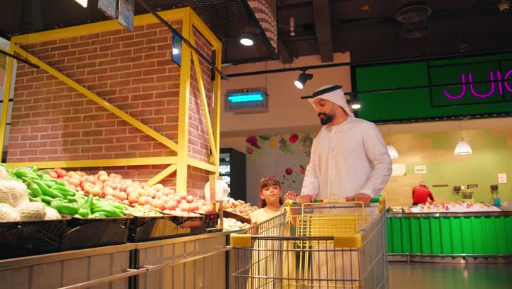 مفهوم قضاء وقت ممتع مع الابناء وشراء الاحتياجات اليومية، رجل عربي خليجي إماراتي يرتدي الكندورة والغترة داخل السوبرماركت يمسك عربة التسوق برفقة ابنته في قسم الخضار والفواكه الطازجة، ايماءات السعادة و السرور، التسوق العائلي داخل المتاجر