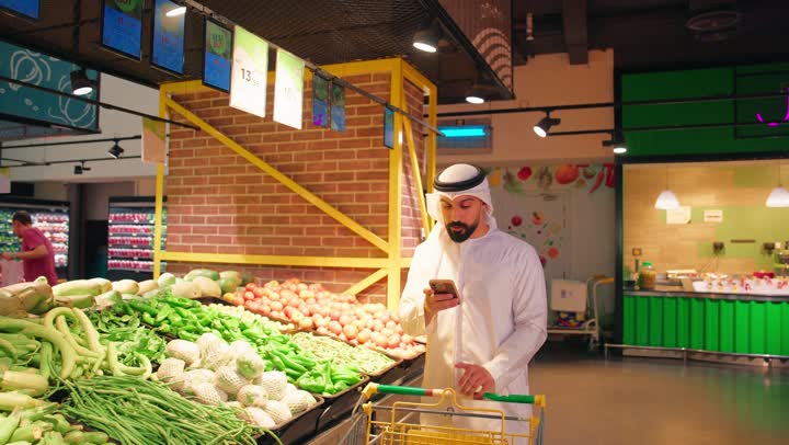 مراجعة قائمة المشتريات أثناء التسوق، مفهوم التسوق الذكي وشراء الاحتياجات اليومية، رجل عربي خليجي إماراتي يرتدي الكندورة والغترة داخل سوبرماركت يجر عربة التسوق في قسم الخضار والفواكه الطازجة يمسك بيده الهاتف المحمول، الاهتمام بجودة المنتجات الغذائية