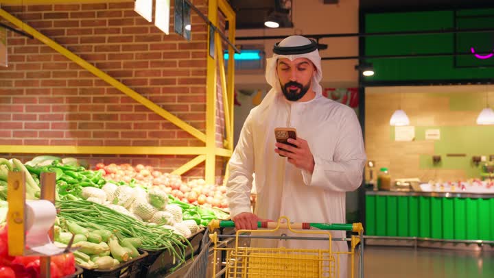 مفهوم التسوق الذكي وشراء الاحتياجات اليومية، رجل عربي خليجي إماراتي يرتدي الكندورة والغترة داخل سوبرماركت يجر العربة في قسم الخضار والفواكه الطازجة يمسك بيده الهاتف المحمول ، مراجعة قائمة المشتريات أثناء التسوق، الاهتمام بجودة المنتجات الغذائية