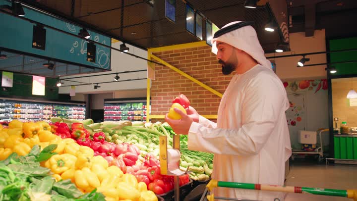 The concept of daily shopping and purchasing household necessities, the focus on the quality of fresh food products, an Arab Gulf Emirati man wearing a kandura and ghutrah inside the supermarket holding two peppers (red and yellow) comparing them before buying.