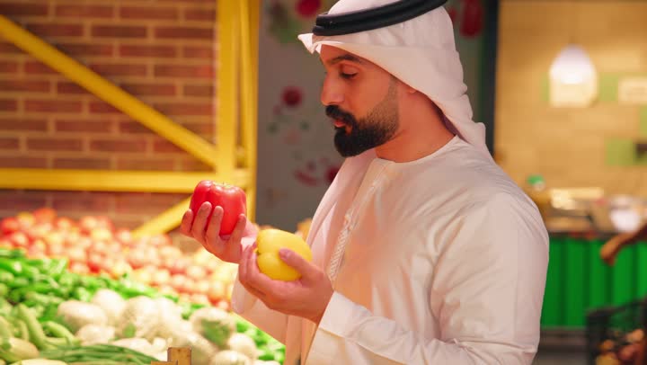 الاهتمام بجودة المنتجات الغذائية الطازجة، تصوير مقرب لرجل عربي خليجي إماراتي يرتدي الكندورة والغترة داخل سوبرماركت  يمسك بيديه حبتين من الفلفل(الأحمر والأصفر) ويفحصهما ويقارن بينهما قبل الشراء، مفهوم التسوق اليومي وشراء الاحتياجات المنزلية