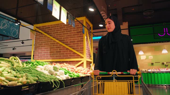 An Arab Gulf Emirati woman wearing a black abaya and hijab inside a supermarket is holding two colorful bell peppers (red and yellow) in her hands and comparing them before purchasing. This reflects the concept of daily shopping and buying household necessities, the focus on the quality of fresh food products, a healthy lifestyle, and proper nutrition.