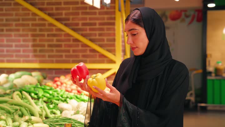شراء المنتجات الطازجة من قسم الخضار في السوبرماركت، امرأة عربية خليجية إماراتية ترتدي العباءة السوداء والحجاب تمسك حبة فلفل أحمر وأصفر وتفحصها بعناية، مفهوم التسوق اليومي