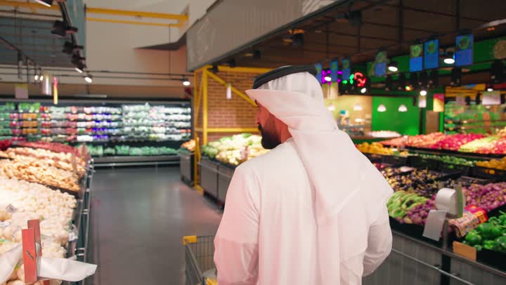 اختيار المنتجات الغذائية بعناية، مفهوم التسوق اليومي وشراء الاحتياجات المنزلية، تصوير من الخلف لرجل عربي خليجي إماراتي يرتدي الكندورة والغترة يتسوق داخل السوبر ماركت يدفع عربة التسوق بين رفوف الخضروات والفواكه الطازجة