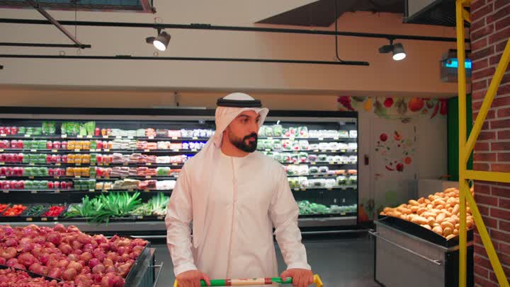 مفهوم التسوق اليومي وشراء الاحتياجات المنزلية، رجل عربي خليجي إماراتي يرتدي الكندورة والغترة يتسوق داخل السوبر ماركت يدفع عربة التسوق بين رفوف الخضروات والفواكه الطازجة، اختيار المنتجات الغذائية بعناية