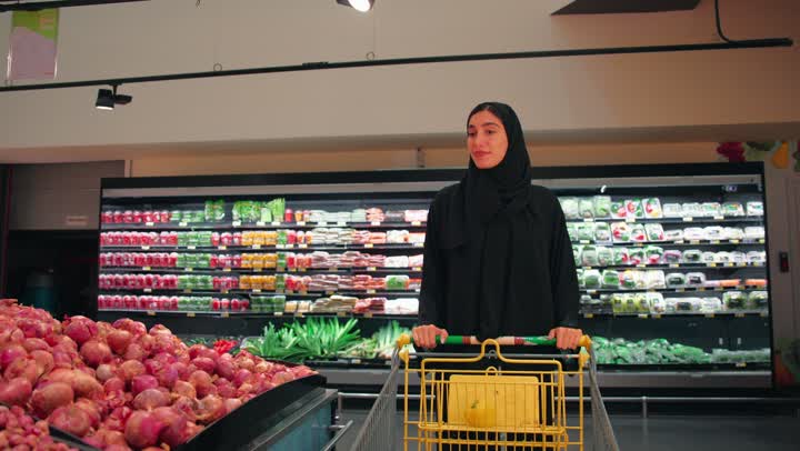 اختيار احتياجات المنزل اليومية، امراة عربية خليجية اماراتية محجبة ترتدي العباءة السوداء تجر العربة تتسوق داخل قسم الخضروات والفواكه في السوبر ماركت الاهتمام بجودة المنتجات الطازجة