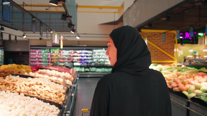 تصوير من الخلف لامرأة عربية خليجية اماراتية محجبة ترتدي العباءة السوداء تجر العربة تتسوق داخل قسم الخضروات والفواكه في السوبر ماركت، اختيار احتياجات المنزل اليومية، الاهتمام بجودة المنتجات الطازجة