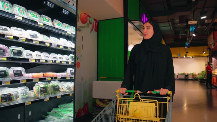 شراء المواد الغذائية والمستلزمات المنزلية، امراة عربية خليجية اماراتية محجبة ترتدي العباءة السوداء تقوم بدفع عربة التسوق داخل السوبر ماركت، التجول بين رفوف المنتجات الغذائية المبردة، الاهتمام بالتغذية الصحية وجودة المنتجات