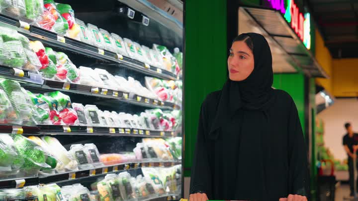 An Arab Gulf Emirati woman wearing a black abaya is selecting food products from the refrigerator shelves inside the supermarket, checking ready-to-eat food packages and canned goods, understanding the concept of daily shopping and providing household necessities, and paying attention to the quality of food products and healthy nutrition.