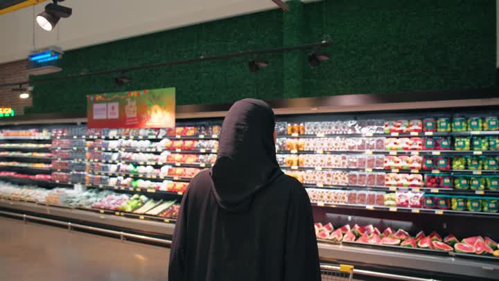 Choosing fresh products and buying daily household needs, the concept of daily shopping and concern for healthy nutrition, a rear view of an Emirati Gulf Arab woman wearing a black abaya, holding a cart and shopping inside the supermarket.