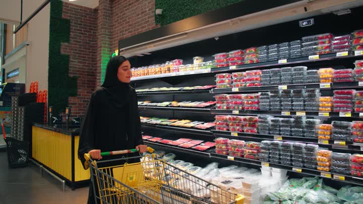 مفهوم التسوق اليومي والاهتمام بالتغذية الصحية، امرأة عربية خليجية اماراتية محجبة ترتدي العباءة السوداء تمسك العربة و تقوم بالتسوق داخل السوبر ماركت، اختيار المنتجات الطازجة وشراء الاحتياجات المنزلية اليومية