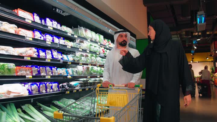 مفهوم التسوق العائلي والتعاون بين الزوجين، رجل عربي خليجي اماراتي يرتدي الزي التقليدي يمسك العربة يقوم بالتسوق داخل السوبر ماركت برفقة زوجتة، اختيار المنتجات الغذائية من رفوف الثلاجات، شراء الاحتياجات المنزلية اليومية، الاهتمام بالتغذية الصحية وجودة الطعام، قضاء وقت ممتع داخل المتجر ، ايماءات السعادة و السرور