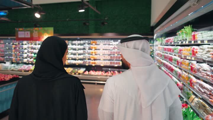 الاهتمام بالتغذية الصحية وجودة الطعام، تصوير من الخلف لرجل وامرأة عربيان خليجيان اماراتيان يرتديان الزي التقليدي يتسوقان داخل السوبر ماركت لاختيار المنتجات الغذائية، التسوق اليومي وشراء الاحتياجات المنزلية، اختيار الخضروات والفواكه الطازجة والمنتجات المعبأة، مفهوم التسوق العائلي
