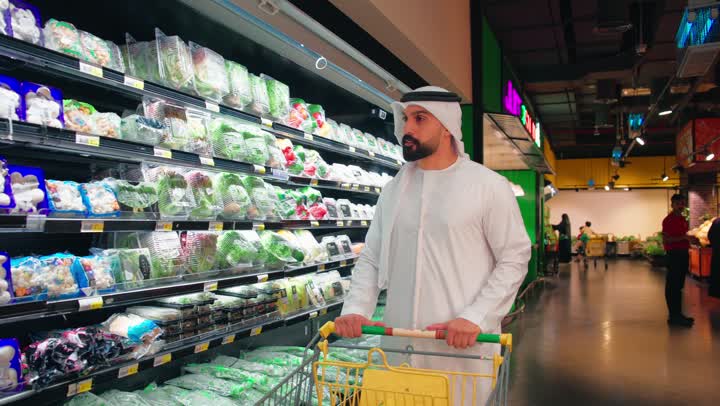 The concept of daily shopping and providing for household needs, buying packaged vegetables and fresh products, an Arab Gulf Emirati man wearing a kandura and ghutrah holding a shopping cart selecting food products from the shelves of refrigerators inside the supermarket, focusing on healthy nutrition and product quality.